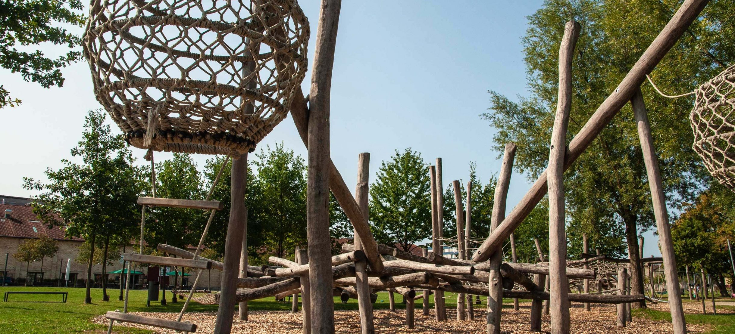 Kinderspielplatz im Eulachpark mit Klettergerüst aus Holzstämmen, einem runden Geflecht mit Strickleiter, umgeben von Bäumen.